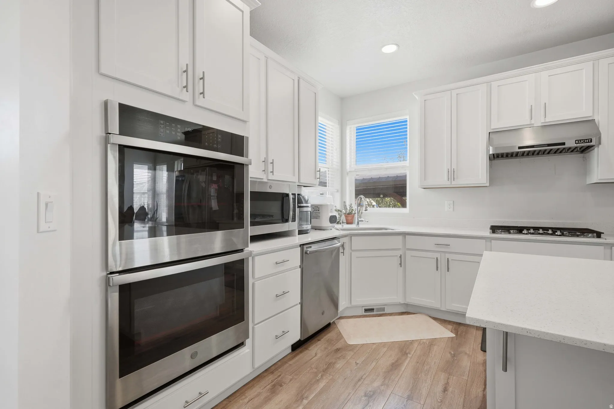 Kitchen featuring white shaker cabinetry, stainless steel double wall ovens, a gas range with a stainless steel vent hood, wood-finish flooring, and light-toned countertops