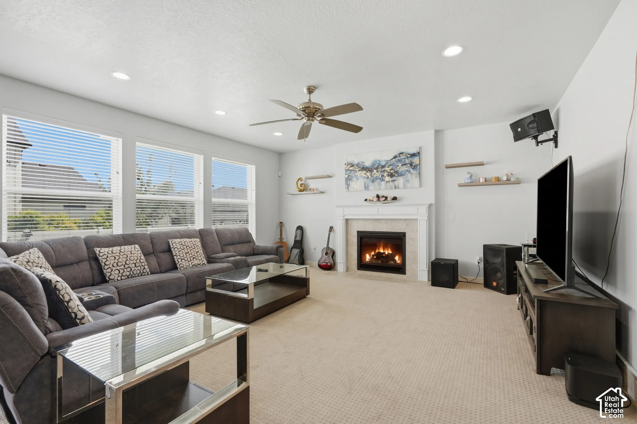 Bright interior space featuring a central fireplace with a white mantel, multiple windows with horizontal blinds, a ceiling fan, recessed lighting, and wall-mounted shelving