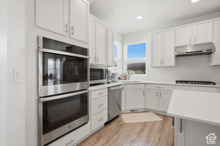 Kitchen featuring white shaker cabinetry, stainless steel double wall ovens, a gas range with a stainless steel vent hood, wood-finish flooring, and light-toned countertops