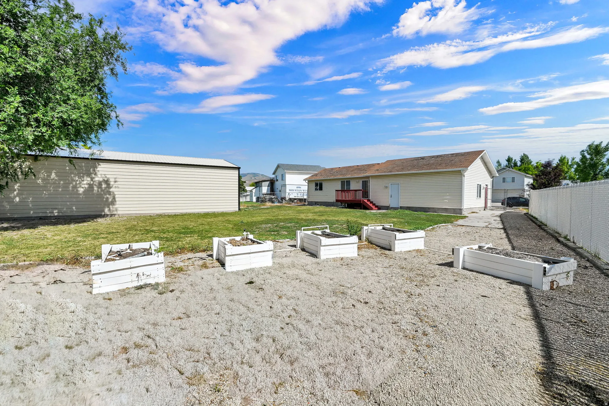 Expansive exterior featuring a cream-colored metal outbuilding, a residential structure with red-painted stairs, and a gravel-covered area with multiple raised garden beds