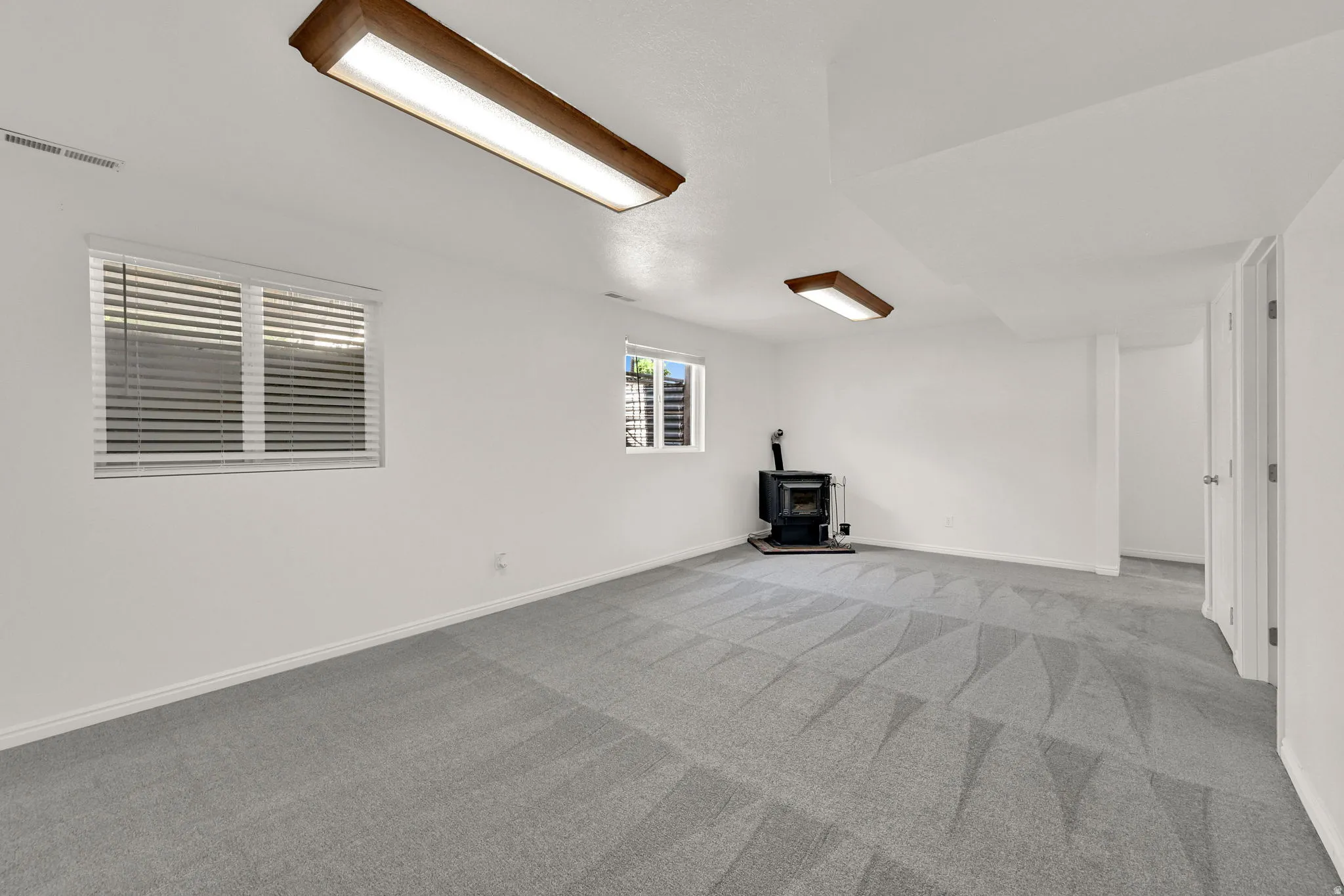 Spacious room featuring light gray patterned carpeting, a freestanding black stove, and two windows with blinds