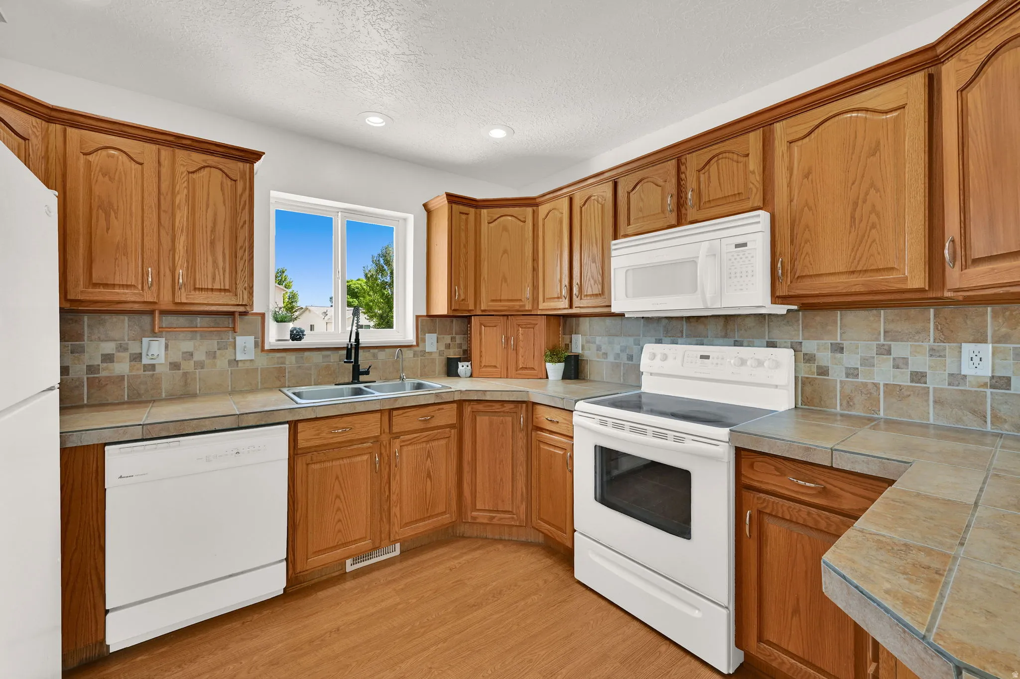 U-shaped kitchen featuring wood-finish cabinetry, tile countertops, a tile backsplash with decorative accents, and a double basin sink with a black-finish faucet
