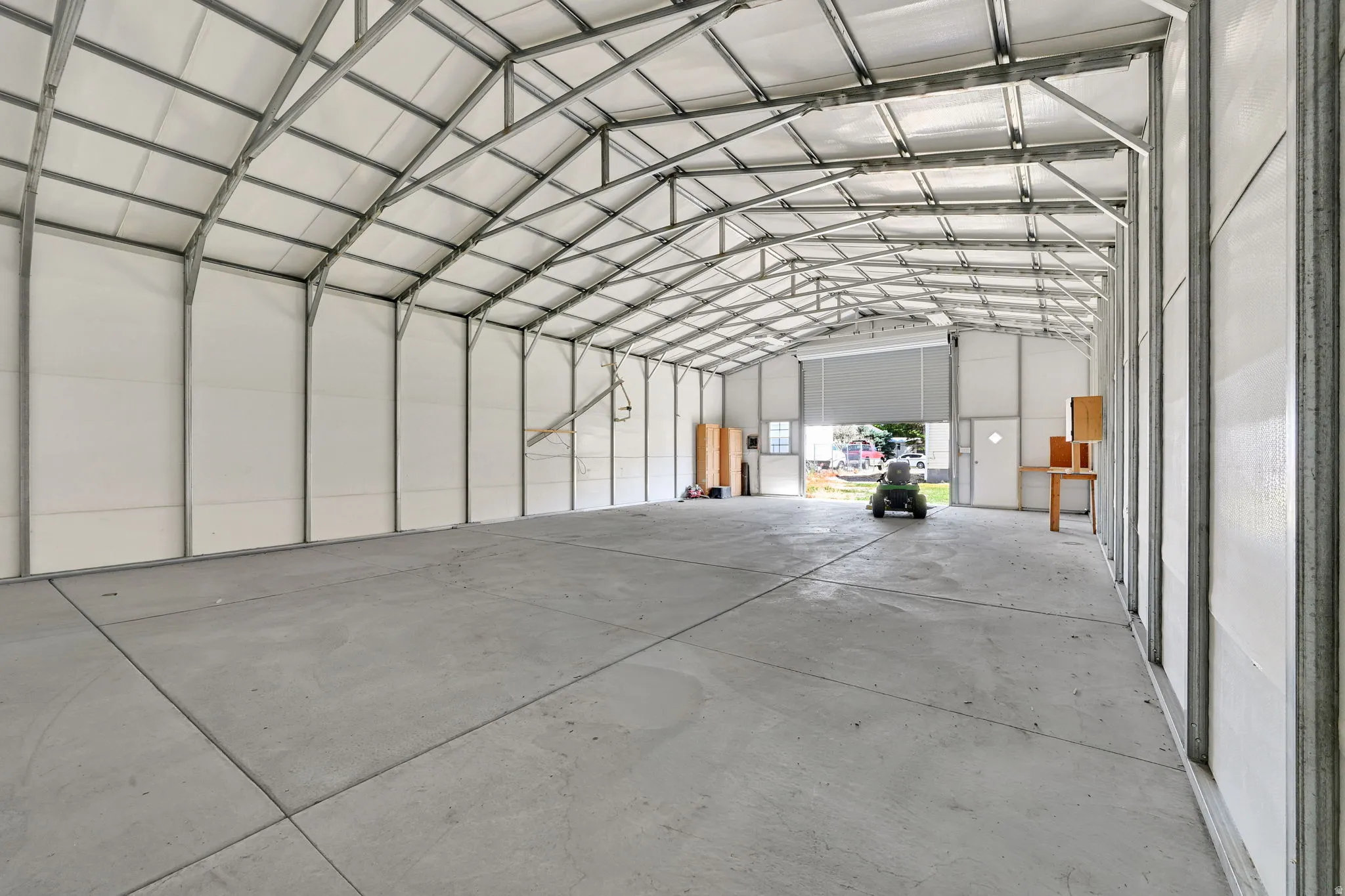 Spacious interior structure featuring an expansive concrete floor, visible metal truss system, arched ceiling, white wall panels, and a large roll-up door