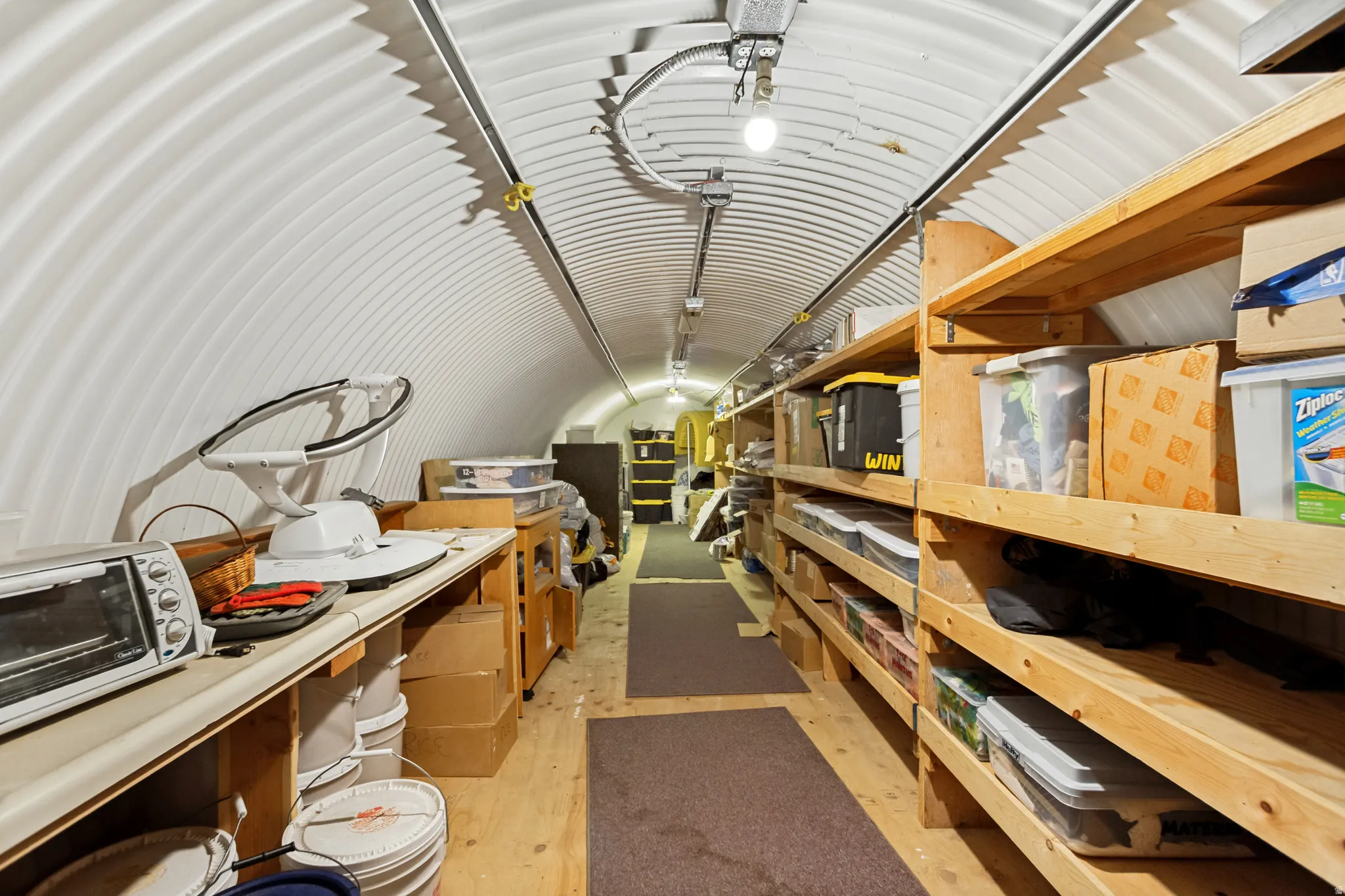 Quonset hut interior featuring a corrugated white arched ceiling, wood-finish flooring, built-in wooden shelving, and track lighting