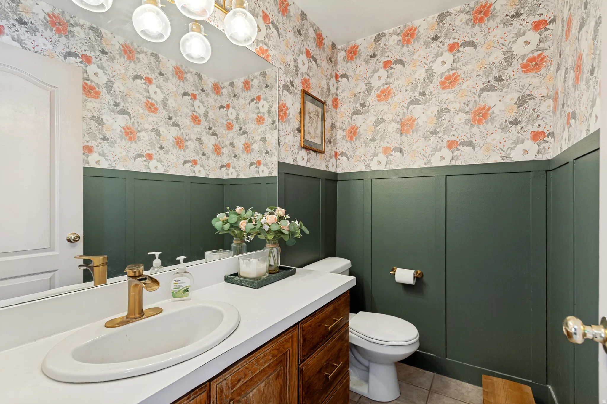 Bathroom featuring board and batten wainscoting with floral wallpaper, a vanity with a white countertop and gold-tone faucet, a large wall-mounted mirror, and tile flooring