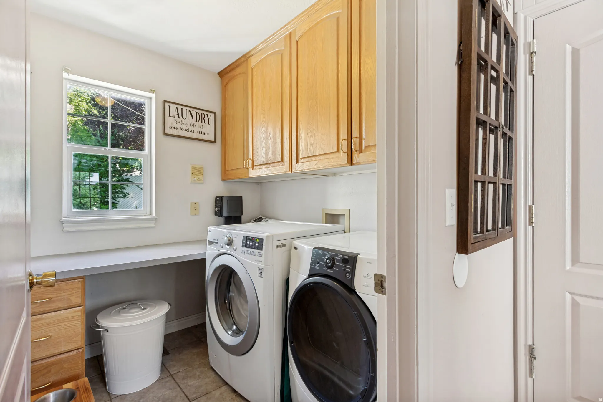 Dedicated laundry area featuring a large window, light wood-finish cabinetry, a built-in countertop, and neutral tile flooring