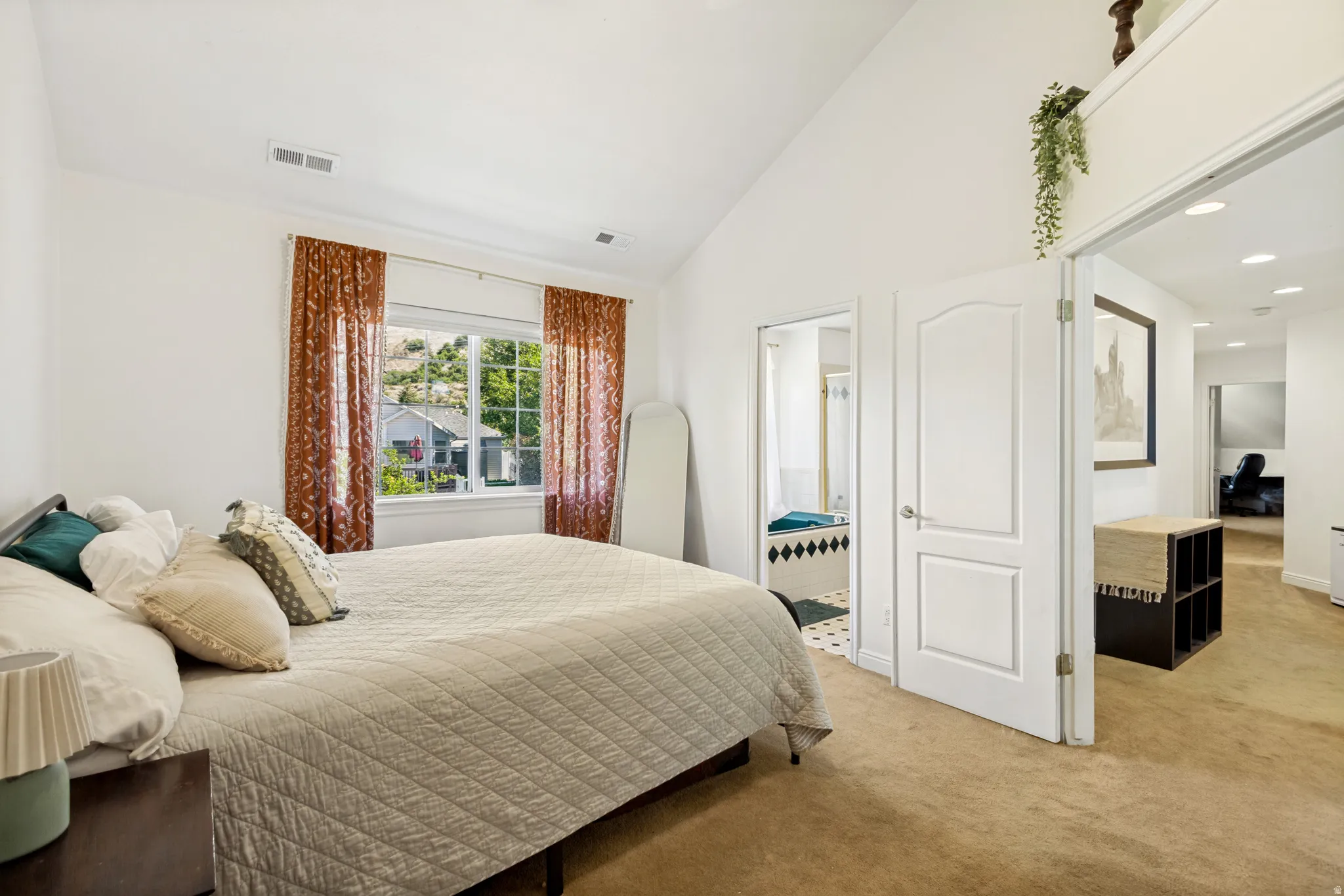 Vaulted ceilings enhance this bedroom space featuring a window with exterior views, carpeted flooring, and a bath area with tiled flooring