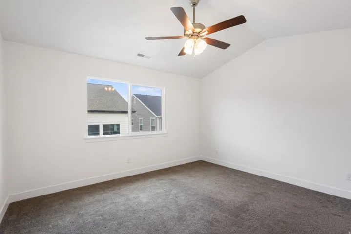 Spacious room featuring vaulted ceilings, a window with exterior views, dark gray carpeting, crisp white walls, and a ceiling fan with integrated lighting