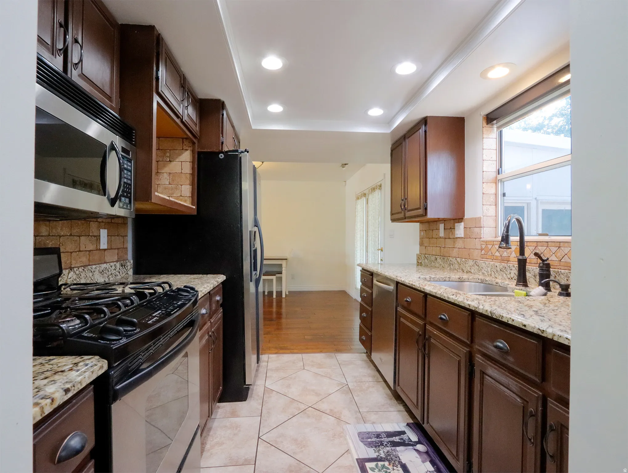 Galley kitchen featuring recessed lighting, dark wood cabinetry, granite countertops, tile backsplash, and stainless steel appliances