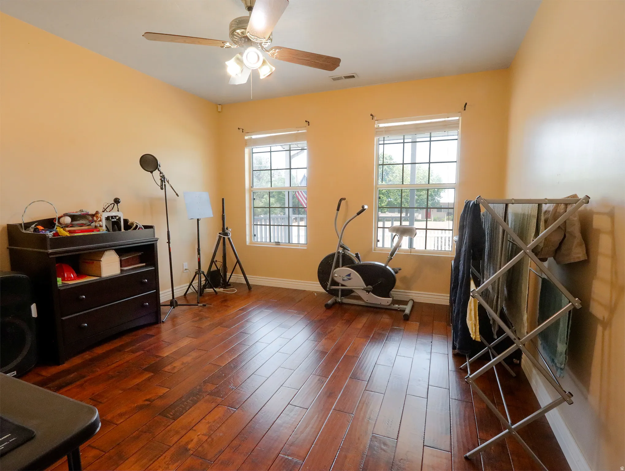 Spacious room featuring rich wood-finish flooring, two large windows with white blinds, and a ceiling fan with integrated lighting