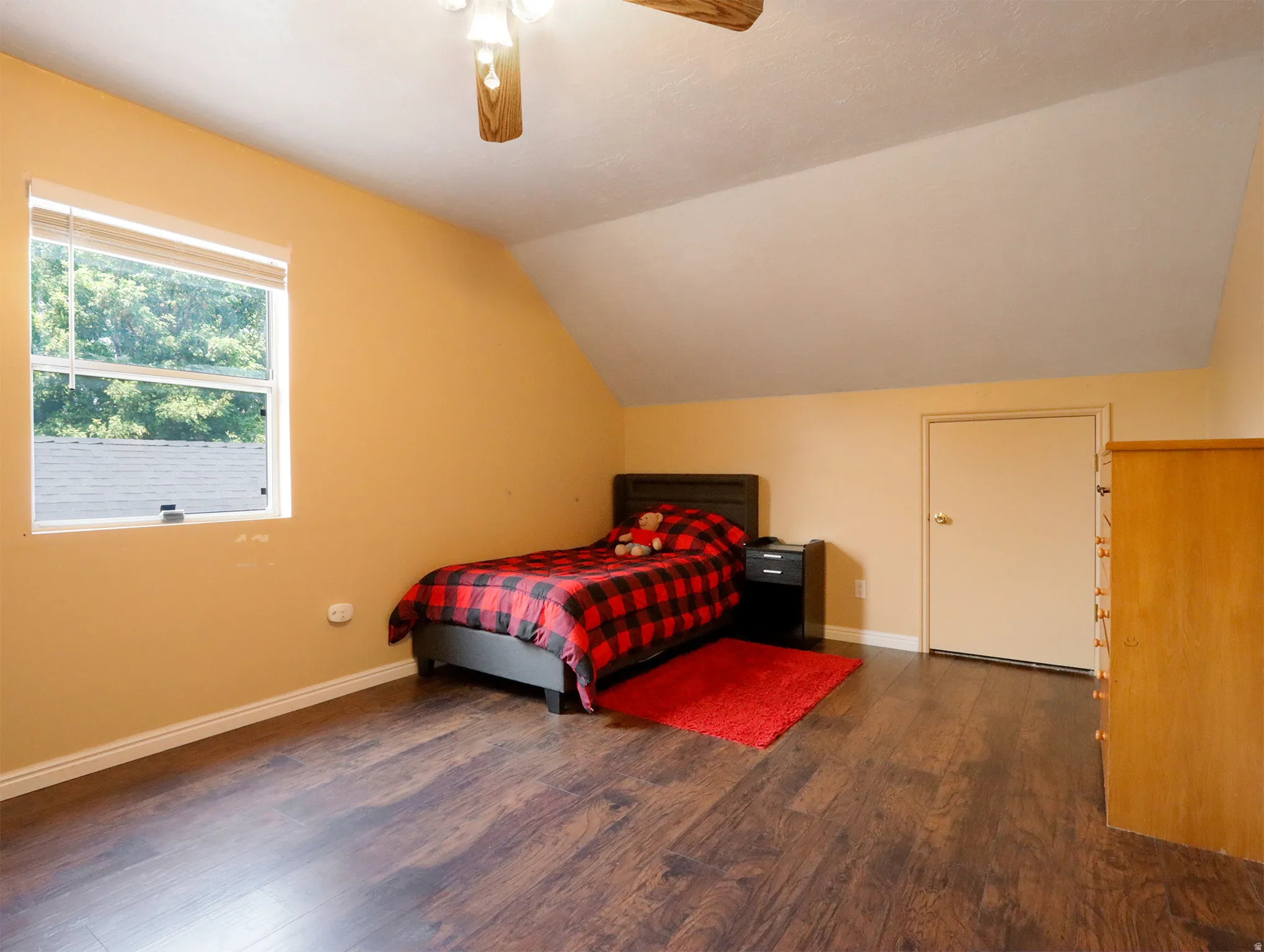 Room featuring rich wood-finish flooring, a single window with blinds, and an angled ceiling