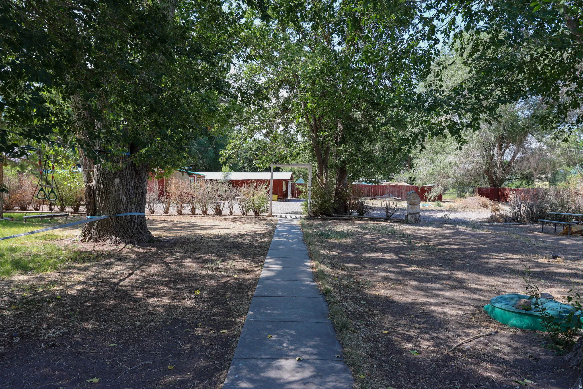 Mature trees provide natural shade and privacy for this property, featuring a concrete pathway and a red fence line