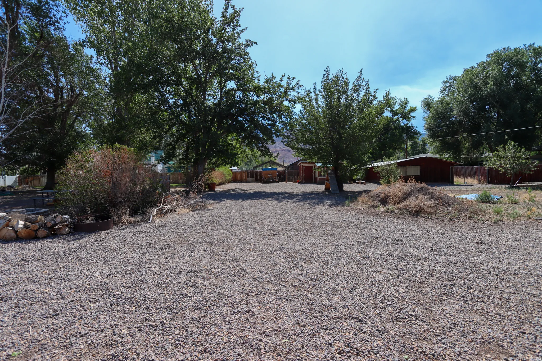 Expansive gravel yard featuring mature trees and a dedicated fire pit area with stone and wood accents