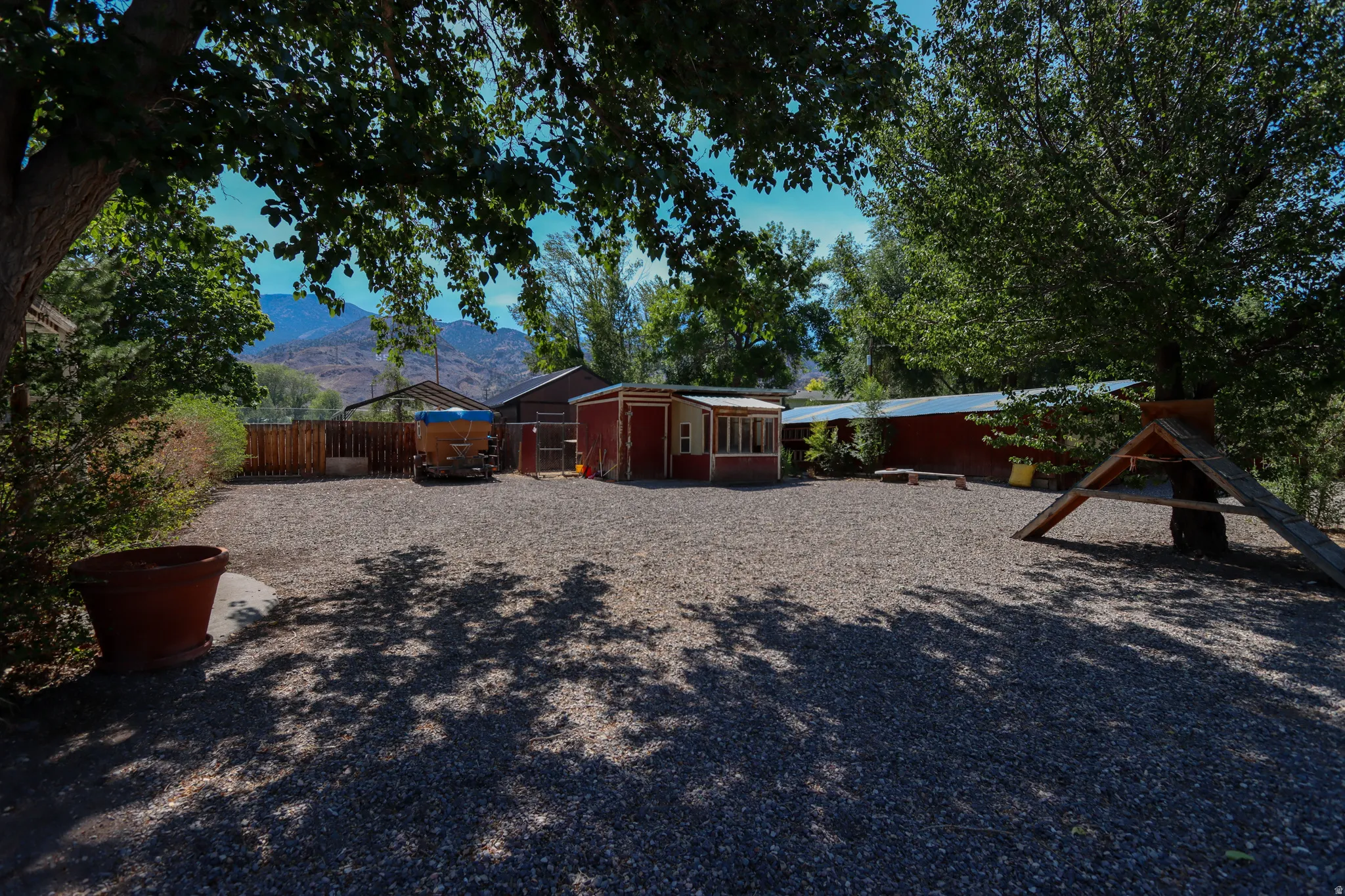 Expansive gravel yard featuring mature trees providing ample shade, mountain backdrop, and multiple outbuildings
