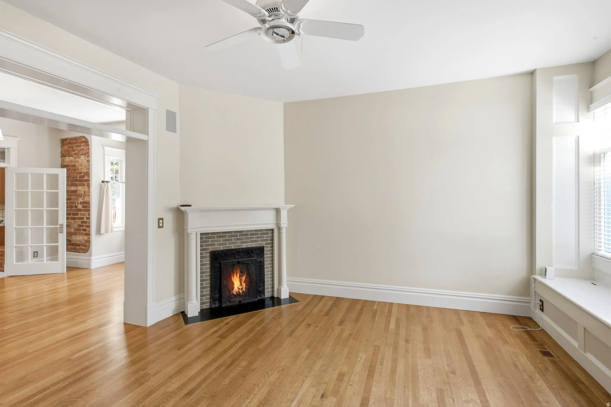 Living space featuring hardwood flooring, a fireplace with a decorative mantel and brick surround, a built-in window bench, a ceiling fan, and French doors opening to an exposed brick wall