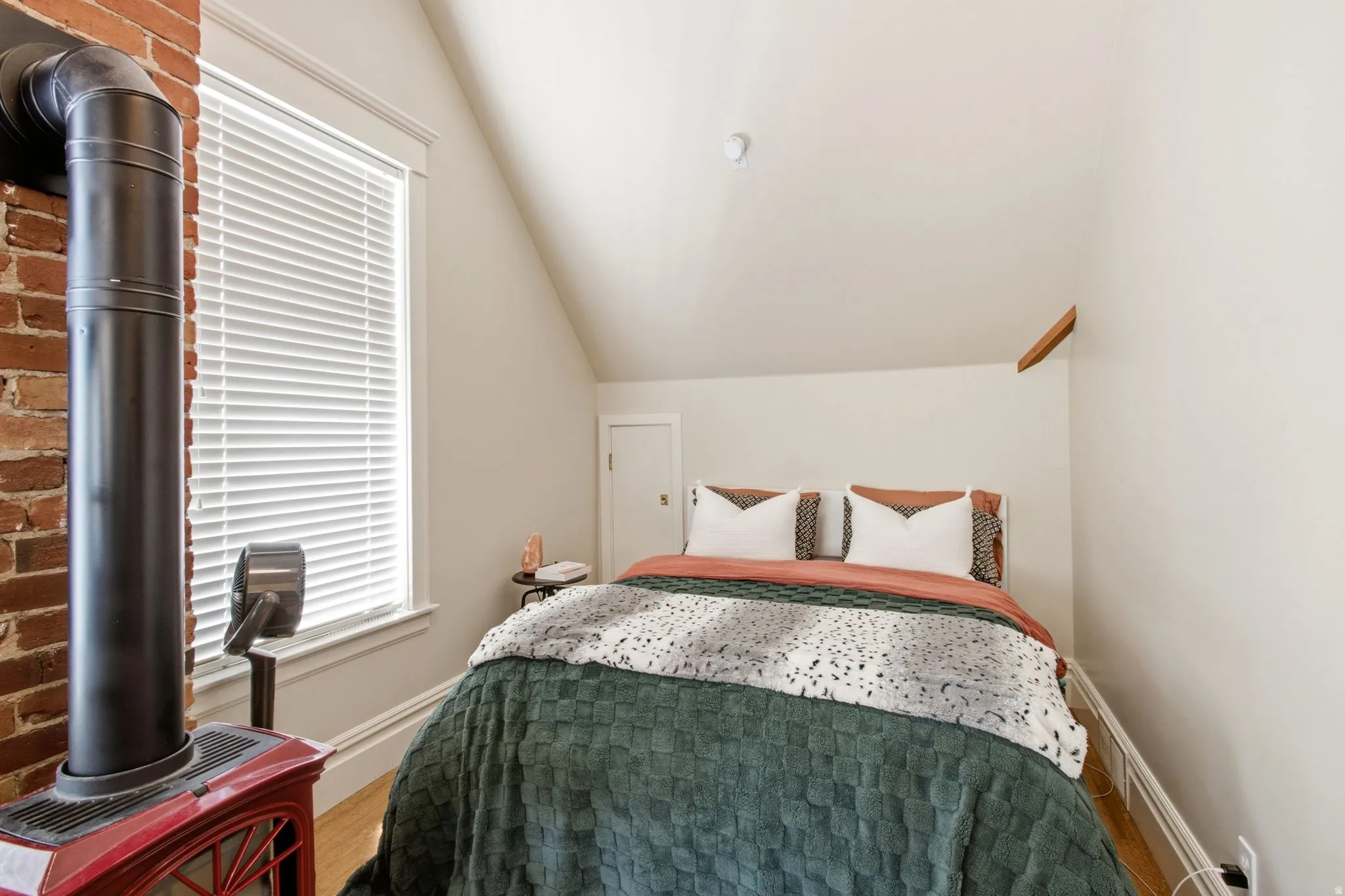 Attic space featuring a red wood-burning stove with exposed brick, a window with blinds, and light wood-finish flooring