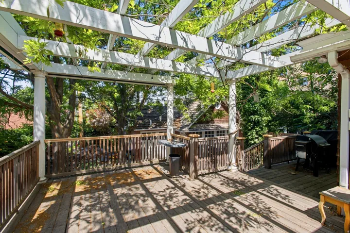 Spacious exterior deck featuring a white pergola with natural vine coverage, wood-finish flooring, and natural wood railing