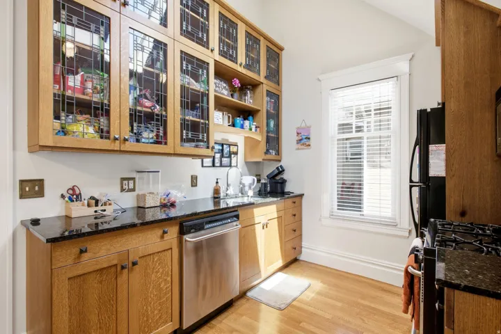 Kitchen featuring wood-finish cabinetry with glass-front upper cabinets, dark stone countertops, stainless steel dishwasher, and wood-finish flooring