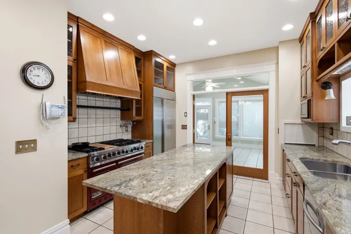 Gourmet kitchen with natural wood cabinetry, light-toned granite countertops, a stainless steel gas range, and a central island with integrated shelving
