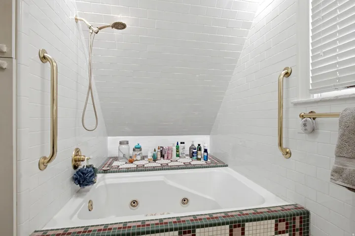 Built-in jetted tub surrounded by white subway tile, featuring a mosaic tile trim, integrated shower fixture, and a window with blinds