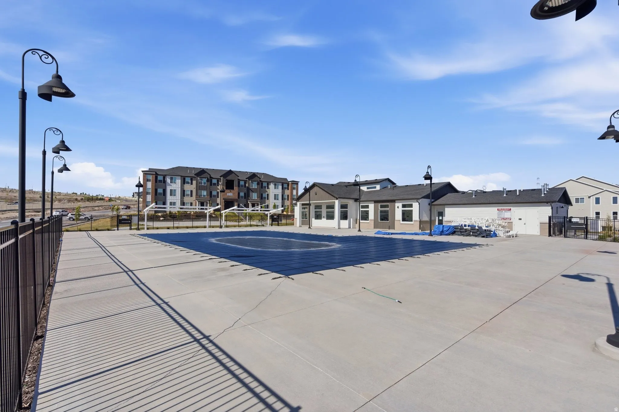 Community pool area with extensive concrete decking, surrounded by a black metal fence, featuring multiple decorative streetlights, and a multi-story residential building in the background