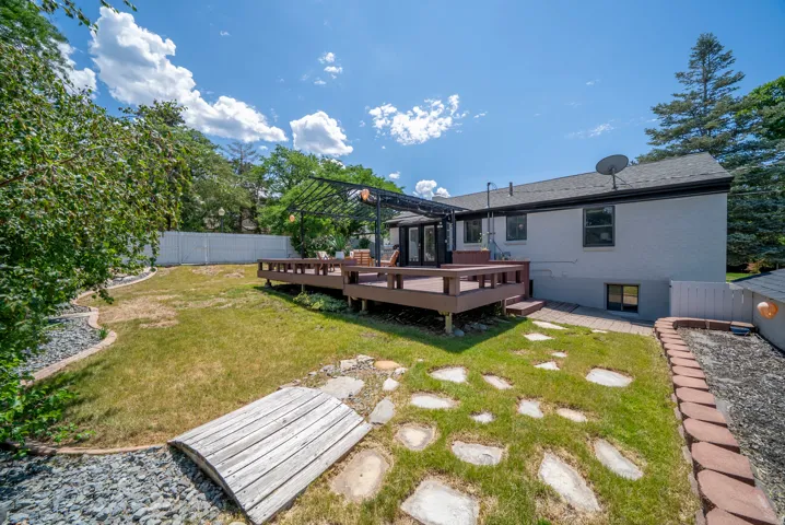 Expansive backyard featuring a multi-level wooden deck with a pergola, a manicured lawn, stone pathway, and a decorative wooden bridge over a rock bed