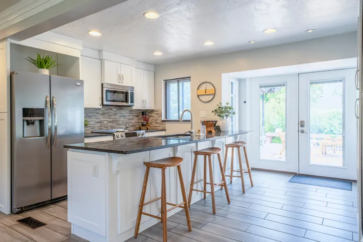 Kitchen featuring white shaker cabinetry, stainless steel appliances, a dark granite countertop island with seating, mosaic tile backsplash, and light wood-finish flooring