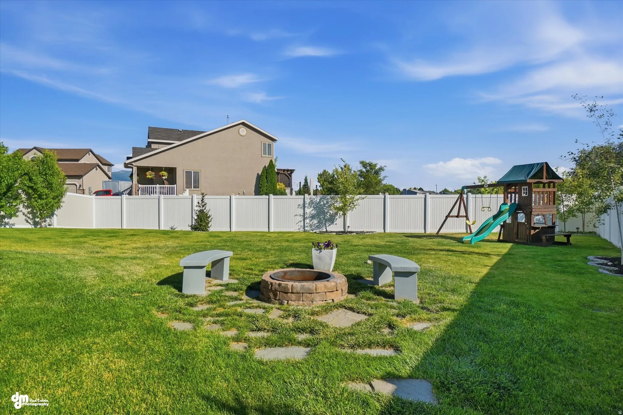 Expansive backyard featuring a stone-paver fire pit with two curved stone benches, a manicured lawn, and a white privacy fence