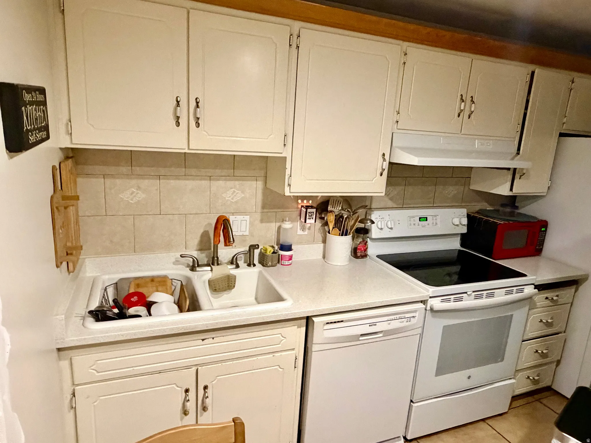 Kitchen featuring cream-colored cabinetry, a tile backsplash, a double basin sink, and white appliances including a stove and dishwasher
