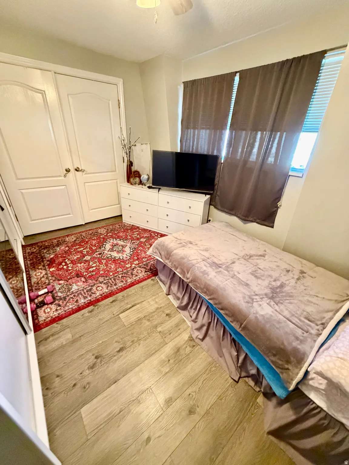 Wood-finish flooring throughout, featuring a double-door closet with arch paneling, a ceiling fan, and window with blinds