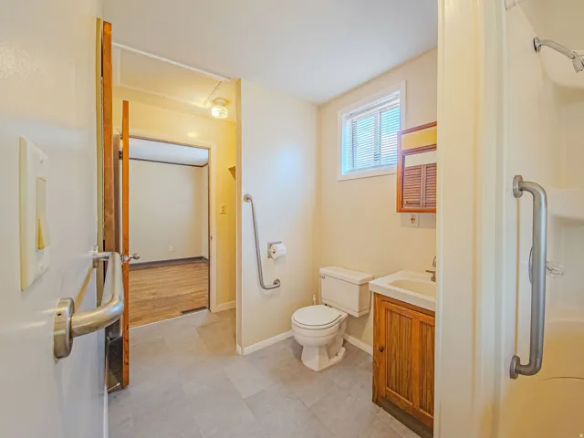 Bathroom featuring grey tile flooring, a window with blinds, a wood-finish vanity with an integrated sink, a toilet, and grab bars