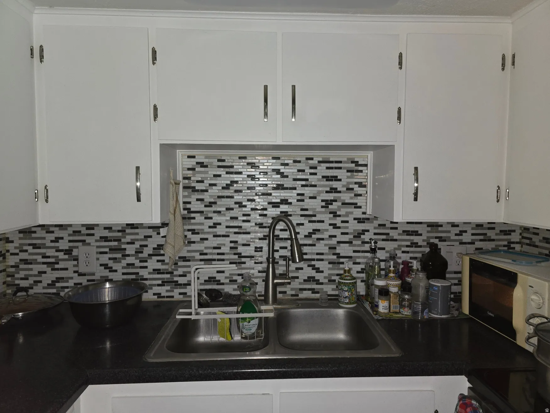 Kitchen featuring a stainless steel double basin sink, mosaic tile backsplash, black countertops, white cabinetry, and brushed nickel hardware