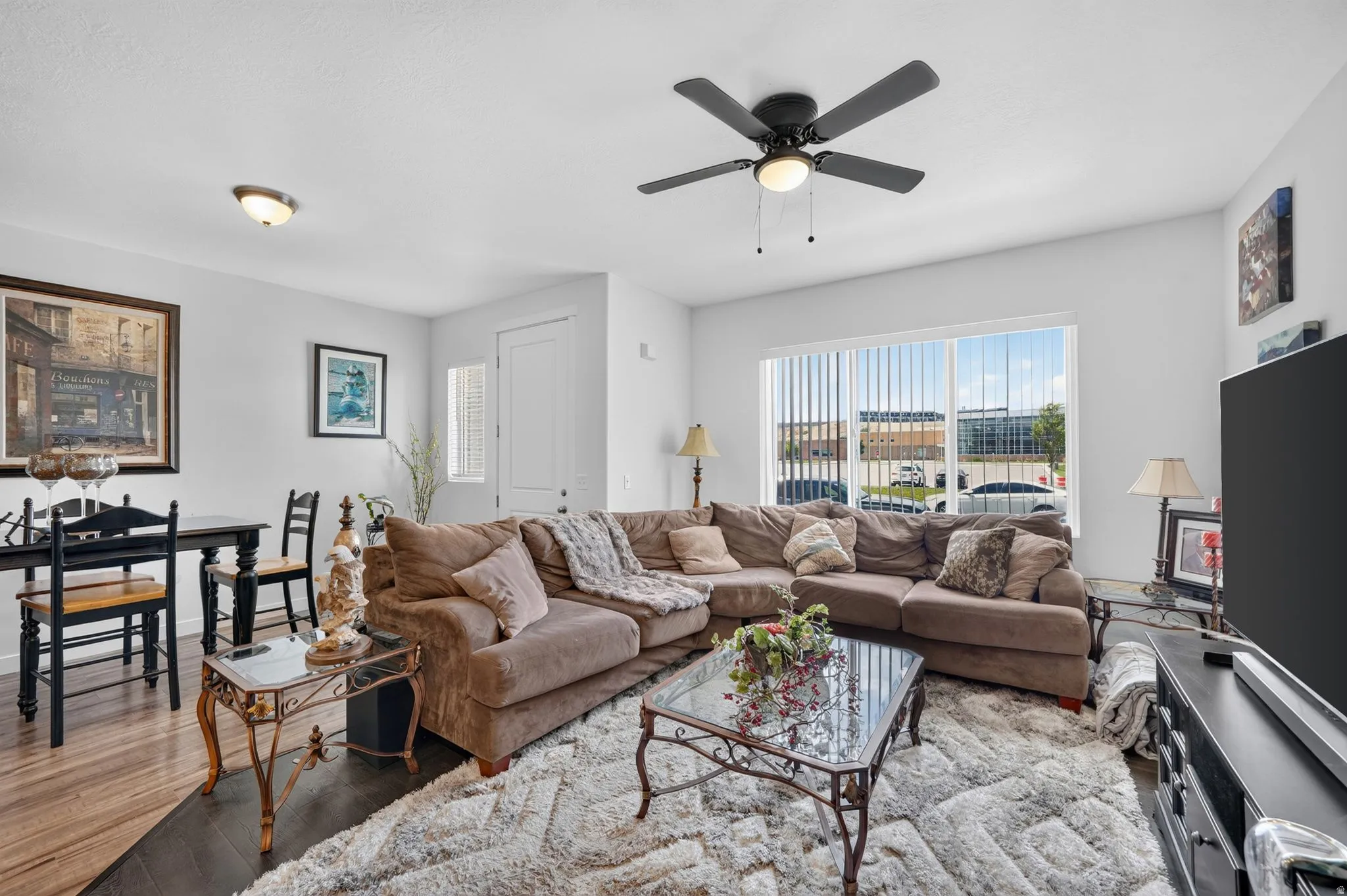 Living area featuring wood-finish flooring, a ceiling fan with integrated lighting, and a large window with vertical blinds