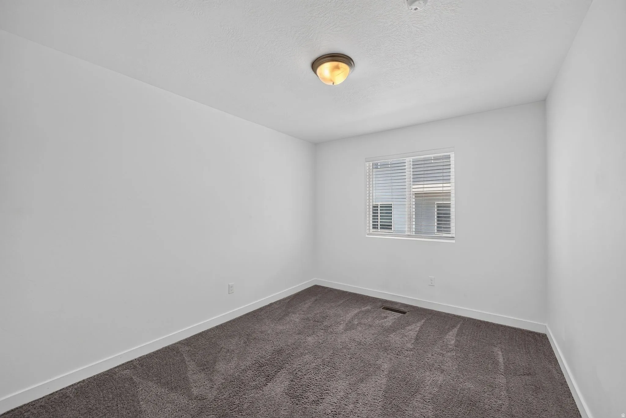 Neutral white walls with a single window, ceiling-mounted light fixture, and dark gray carpeting
