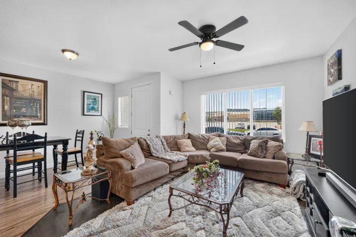 Living area featuring wood-finish flooring, a ceiling fan with integrated lighting, and a large window with vertical blinds