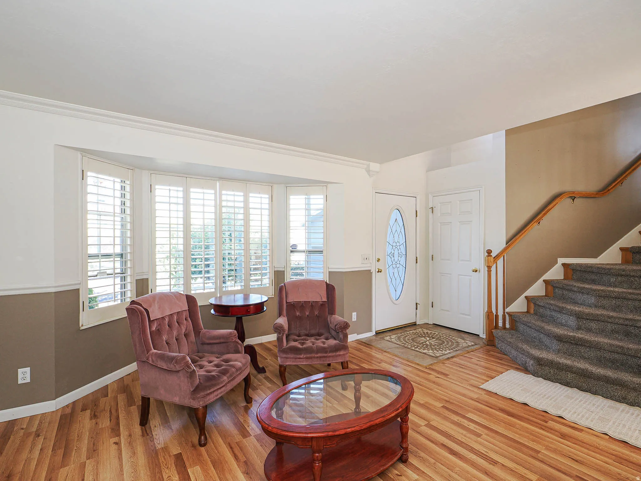 Bay window with plantation shutters, wood-finish flooring, staircase with carpeted treads and wooden handrail, and a front entry door with decorative glass inlay