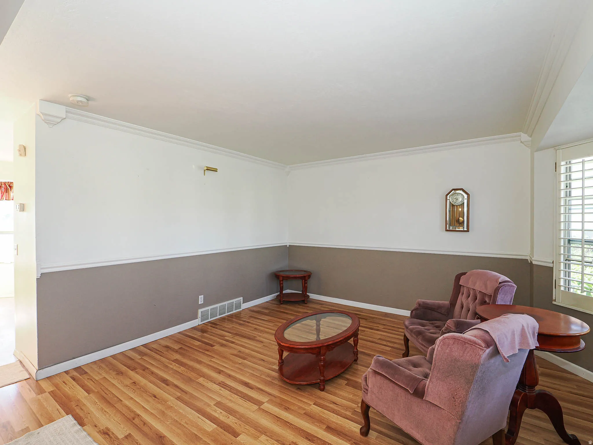 Spacious room featuring crown molding, two-tone wall paint, wood-finish flooring, and white plantation shutters on a bay window