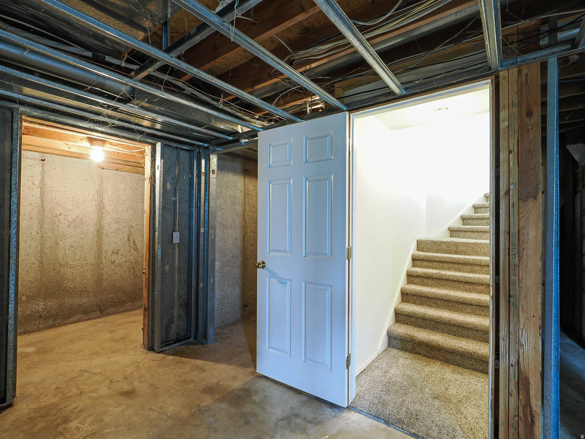 Unfinished basement space featuring exposed ceiling joists and structural beams, concrete flooring, and a white paneled door opening to a carpeted staircase