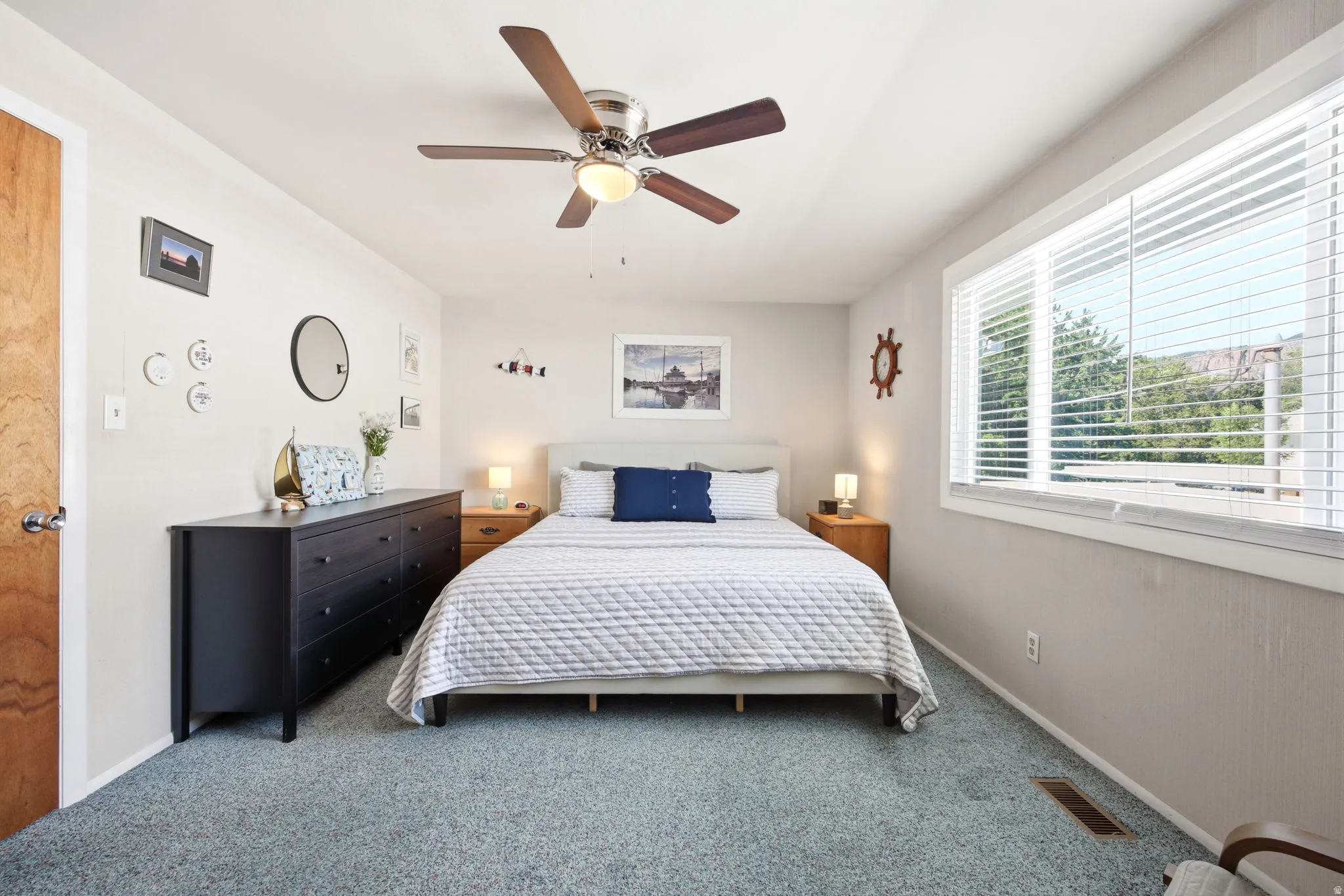 Carpeted bedroom featuring a large window with blinds, a ceiling fan with light, and a wood-finish door