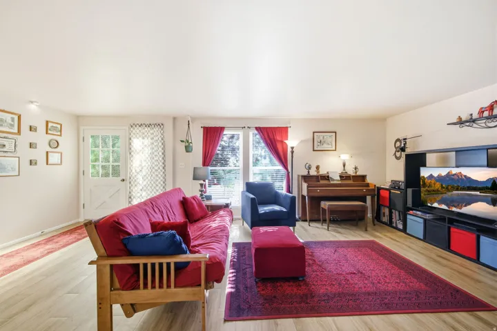 Spacious living area featuring light wood-finish flooring, a white glass-paneled door, and large windows with red drapes providing natural illumination