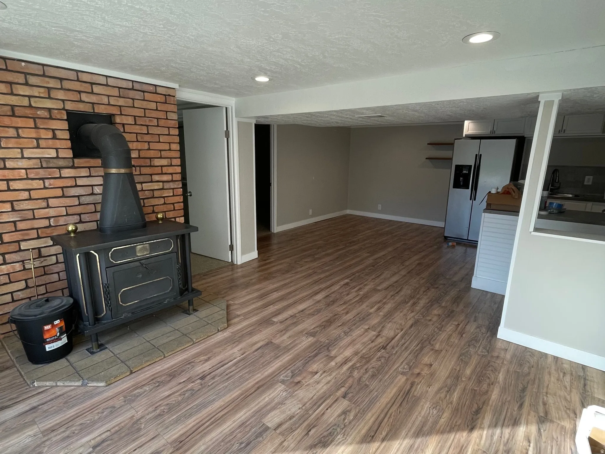 Open-concept living area featuring wood-finish flooring, recessed lighting, and a brick accent wall with a wood-burning stove