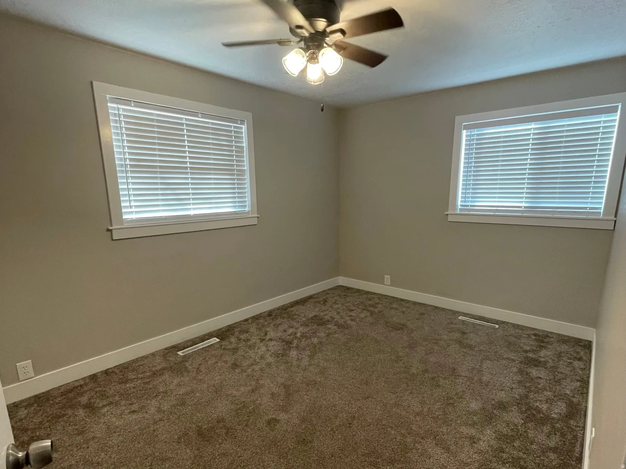 Carpeted room featuring a ceiling fan with light fixture, two windows with blinds, neutral wall paint, and white baseboards