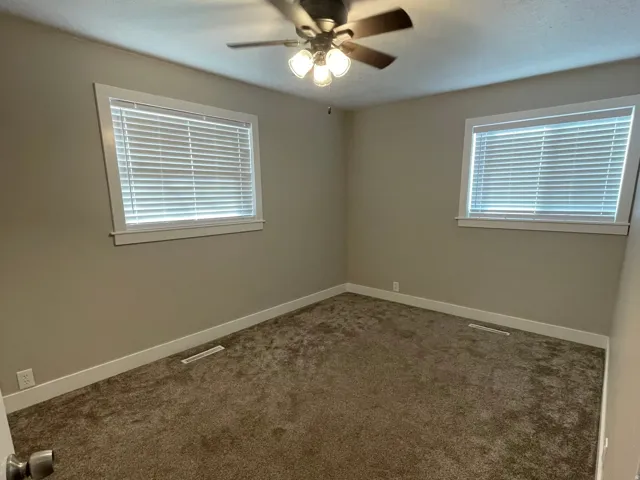 Carpeted room featuring a ceiling fan with light fixture, two windows with blinds, neutral wall paint, and white baseboards