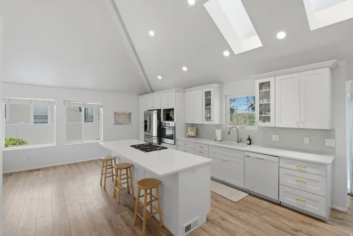 Kitchen featuring a vaulted ceiling with skylights, wood-finish flooring, white cabinetry, a large center island with a built-in cooktop, and stainless steel appliances