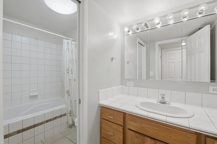 Bathroom featuring a built-in bathtub with white tile surround, a wood-finish vanity, a white tile countertop, and a mirrored medicine cabinet with overhead lighting