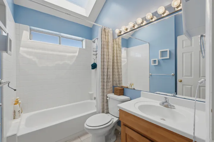 Bathroom featuring a skylight, white tile shower surround, window above the shower, wood-finish vanity, and a large mirror with integrated lighting