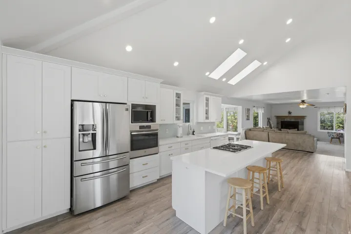 Open-concept kitchen featuring white shaker cabinetry, stainless steel appliances, a center island with a built-in cooktop, light-toned wood-finish flooring, and vaulted ceilings with skylights
