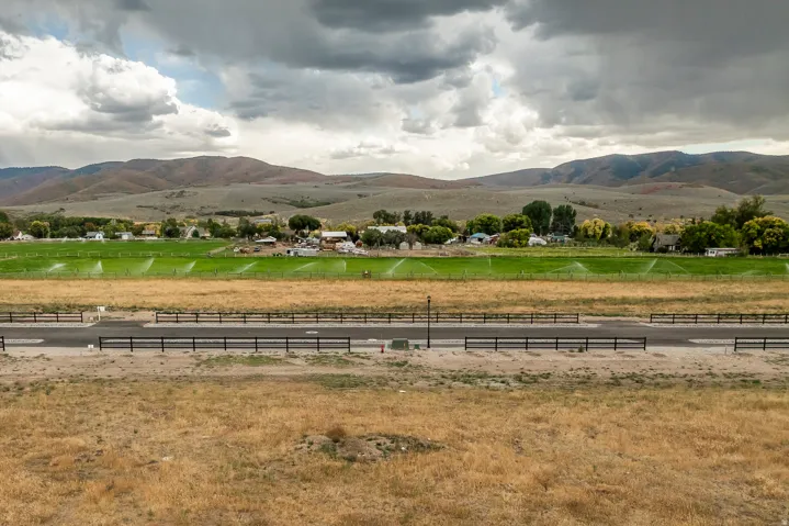 Expansive property featuring a paved road, dark fencing, agricultural land with active sprinklers, and distant residential structures against a backdrop of rolling hills