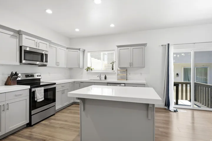 Modern kitchen featuring light gray cabinetry, white countertops, wood-finish flooring, stainless steel appliances, and a sliding glass door to an outdoor deck