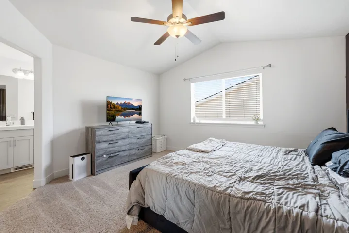 Spacious room featuring vaulted ceilings, carpet flooring, a ceiling fan with integrated lighting, and a private doorway to a bathroom with a vanity and built-in cabinetry